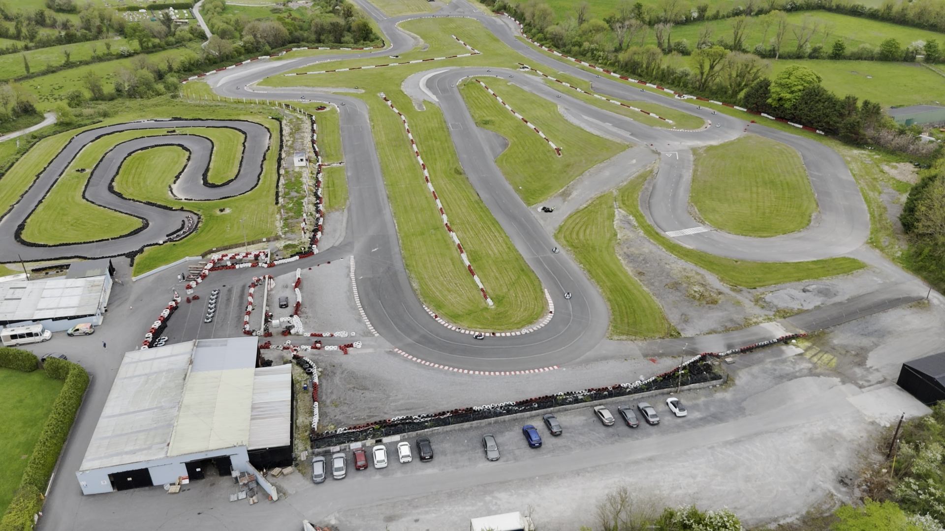 Aerial view of a go-kart racing track with winding paved lanes, green grassy areas, parked cars, and an RSA Rallysport Association building near the entrance, all surrounded by trees and fields.