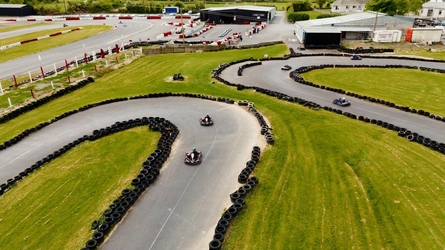 Aerial view of a karting track with several people driving go-karts around winding turns, surrounded by grass and black tire barriers. Buildings and parked cars are visible in the background.